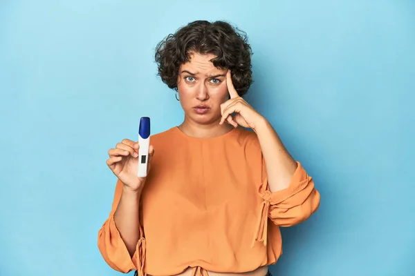 Young woman holding pregnancy test, studio background pointing temple with finger, thinking, focused on a task.