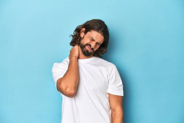 Beyaz gömlekli sakallı bir adam, mavi arka planda stresten dolayı boynu ağrıyor, masaj yapıyor ve elle dokunuyor..