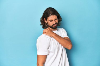Beyaz gömlekli sakallı adam, mavi arka planda omuz ağrısı var..