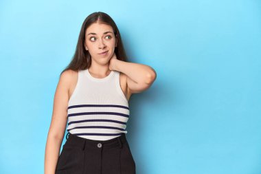 Çizgili bluzlu bir kadın mavi bir stüdyoda poz veriyor kafasının arkasına dokunuyor, düşünüyor ve bir seçim yapıyor..