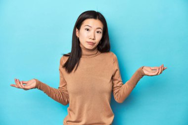 Kahverengi balıkçı yakalı genç Asyalı kadın, sorgulama jestinde şüphe ediyor ve omuz silkiyor..