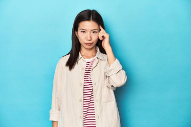 Katmanlı tişörtlü ve çizgili tişörtlü Asyalı bir kadın şakağı parmağıyla gösteriyor, düşünüyor ve bir göreve odaklanıyor..