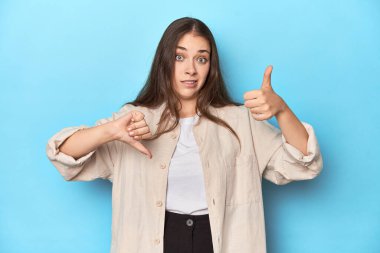 Mavi arkaplanda üst üste giyinmiş zarif genç bir kadın baş parmağını kaldırıyor, zor bir seçim.