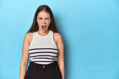 Çizgili bluzlu bir kadın mavi bir stüdyo arkaplanında poz veriyor çok kızgın, öfke dolu bir konsept bağırıyor..