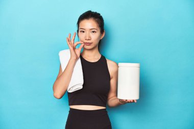 Protein şişesi olan genç Asyalı sporcu, spor salonu düzeneği, parmakları dudaklarında bir sır saklayan.