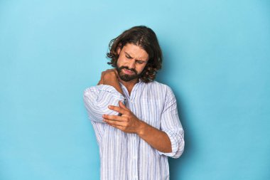 Mavi çizgili tişörtlü sakallı bir adam, mavi stüdyoda dirsek masajı yapıyor, kötü bir hareketin ardından acı çekiyor..