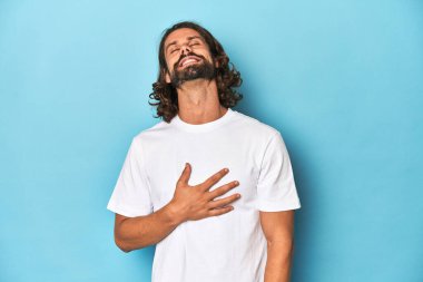 Beyaz tişörtlü sakallı adam, mavi arka planda kahkahalar atıyor elini göğsünde tutuyor..