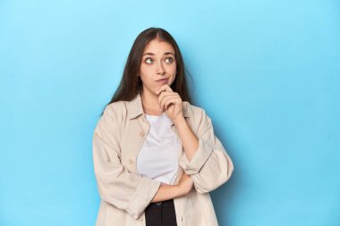 Stylish young woman in an overshirt on a blue background thinking and looking up, being reflective, contemplating, having a fantasy. clipart
