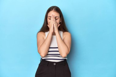 Çizgili bluzlu bir kadın mavi bir stüdyoda poz veriyor. El ele tutuşup, ağza yakın dua ediyor. Kendine güveniyor..