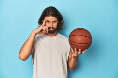 Mavi stüdyoda basketbolcu sakallı bir adam şakağı parmağıyla işaret ediyor, düşünüyor ve bir göreve odaklanıyor..