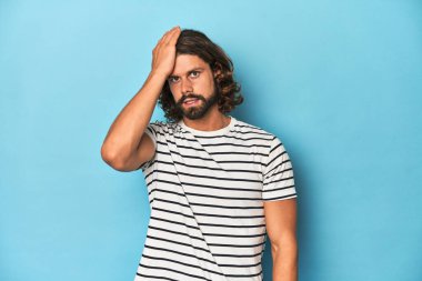 Çizgili gömlekli sakallı bir adam, mavi arka planda bir şeyi unutuyor, alnına avuç içi ve kapalı gözlerle vuruyor..