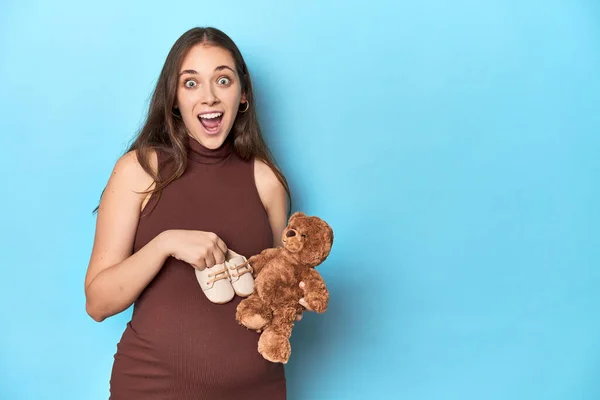 Pregnant woman holding a baby toy and shoes on blue studio.