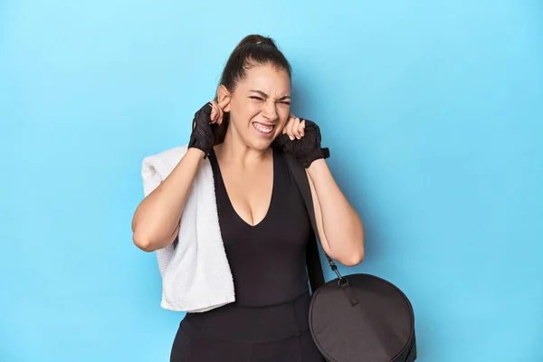 Young woman ready for gym with essentials on blue backdrop covering ears with hands.