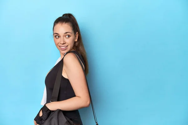 Young woman ready for gym with essentials on blue backdrop looks aside smiling, cheerful and pleasant.
