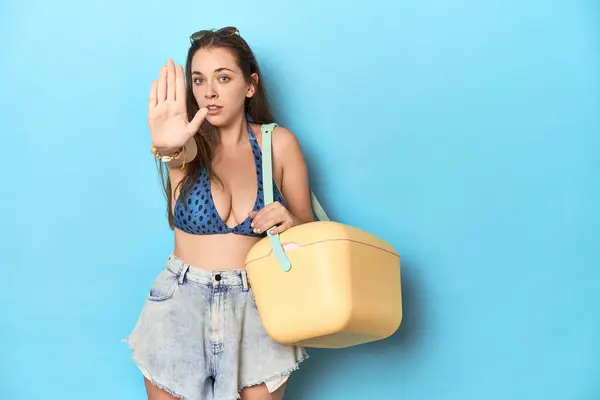 ポータブルビーチクーラー付きのビキニの女性 ストップサインを示す外側の手で立っている青いスタジオ — ストック写真