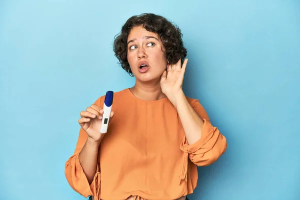 Young woman holding pregnancy test, studio background trying to listening a gossip.