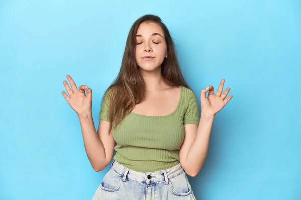 Young Caucasian woman in a green top on a blue backdrop relaxes after hard working day, she is performing yoga.