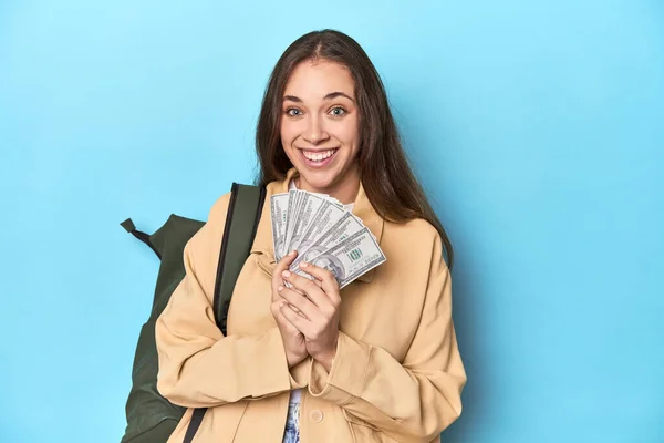 Traveling woman with backpack holding dollar bills on blue.