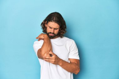 Beyaz tişörtlü sakallı adam, mavi arka plan masajcısı dirsek, kötü bir hareketin ardından acı çekiyor..