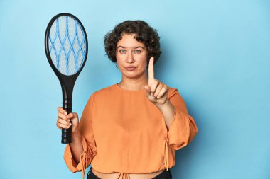 Elektrikli sivrisinek raketi olan genç bir kadın. Elinde elektrikli sivrisinek raketi olan genç bir kadın. Parmağıyla bir numarayı gösteriyor..