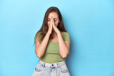 Mavi bir arka planda yeşil bluzlu genç beyaz kadın el ele tutuşup dua ediyor, kendinden emin..