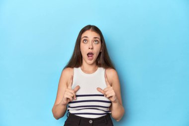 Çizgili bluzlu bir kadın mavi bir stüdyo arkaplanında poz veriyor üst tarafı açık ağzıyla gösteriyor..