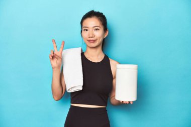 Protein şişesi olan genç Asyalı sporcu, spor salonu düzeneği, parmaklarıyla iki numarayı gösteriyor..