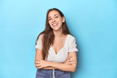 Mavi bir stüdyo arka planında beyaz bluzlu şık genç bir kadın gülüyor ve eğleniyor..