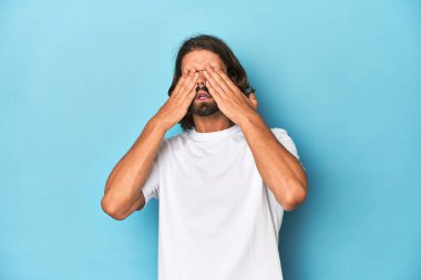 Beyaz tişörtlü sakallı bir adam, mavi arka plan, elleri ve gözleri kapalı..