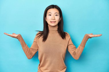 Kahverengi balıkçı yakalı genç Asyalı kadın. Kafası karışmış ve omuz silkiyor. Kopya alanını tutmak için..