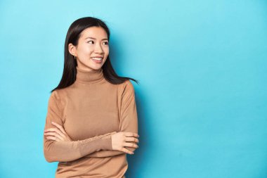 Kahverengi balıkçı yakalı genç Asyalı kadın, kollarını kavuşturarak gülümsüyor..