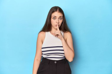 Çizgili bluzlu bir kadın mavi bir stüdyoda poz veriyor ya sır saklıyor ya da sessizlik istiyor..