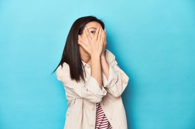 Katmanlı tişörtlü, çizgili tişörtlü Asyalı bir kadın parmaklarının arasından korkmuş ve gergin bir şekilde göz kırpıyor..