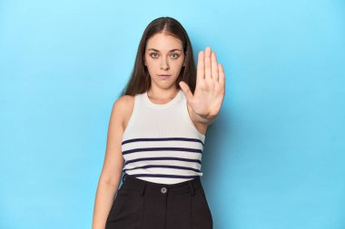 Çizgili bluzlu bir kadın mavi bir stüdyo arkaplanında poz veriyor. Elini uzatıp dur işareti gösteriyor..