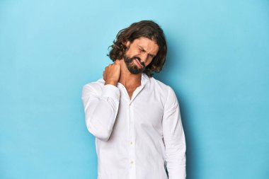 Beyaz tişörtlü sakallı bir adam, mavi arka planda sabit yaşam tarzından dolayı boyun ağrısı çekiyor..