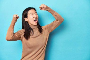 Kahverengi balıkçı yakalı genç Asyalı kadın, zaferden sonra yumruğunu kaldırıyor, kazanan konsept.