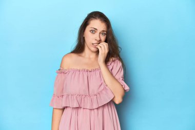Mavi bir stüdyo stüdyosunda pembe elbise giyen genç bir kadın tırnak yiyor, gergin ve çok endişeli..