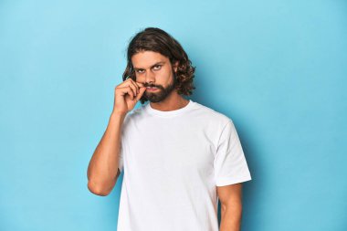 Beyaz gömlekli sakallı bir adam, mavi arka planda dudakları dudaklarında bir sır saklıyor..