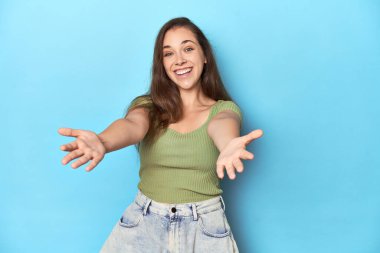 Young Caucasian woman in a green top on a blue backdrop showing a welcome expression. clipart