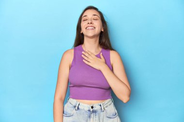 Mor bluzlu, mavi arka planda şık genç bir kadın elini göğsünde tutarak kahkahalar atıyor..