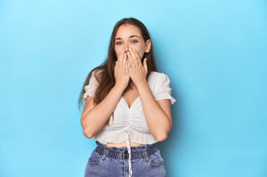 Mavi bir stüdyo arka planında beyaz bluz giymiş zarif genç bir kadın. Bir şeylere gülüyor, elleriyle ağzını kapatıyor..
