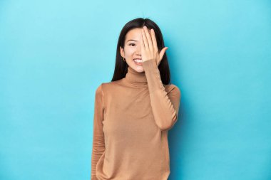 Kahverengi balıkçı yakalı genç Asyalı kadın, yüzünün yarısını palmiye ile örterek eğleniyor..