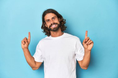 Beyaz tişörtlü sakallı bir adam, mavi arka plan iki ön parmağıyla boş bir alan gösteriyor..
