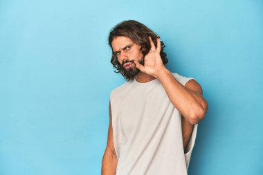 Askılı tişörtlü sakallı bir adam, mavi arka planda bir dedikoduyu dinlemeye çalışıyor..