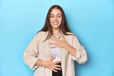 Mavi arka planda tişörtlü şık genç bir kadın mutlu bir şekilde gülüyor ve ellerini karnının üstünde tutarak eğleniyor..