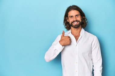 Beyaz gömlekli sakallı adam, mavi arka planda gülümseyen ve baş parmağını kaldıran.