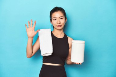 Protein şişeli genç Asyalı sporcu, spor salonu düzeneği, parmaklarıyla beş numarayı gösteren neşeli gülümseme..