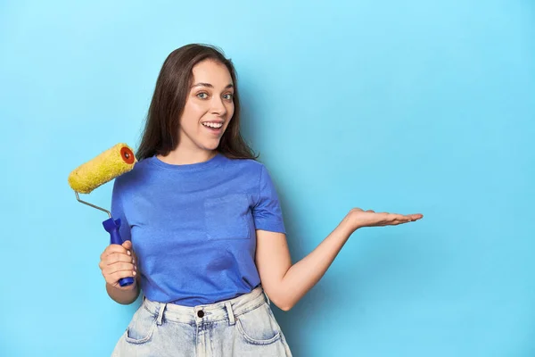 青い背景に黄色いペイントローラーを塗った若い女性は 手のひらにコピースペースを表示し ウエストに別の手を保持 — ストック写真