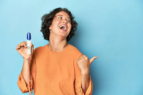 Young woman holding pregnancy test, studio background points with thumb finger away, laughing and carefree.