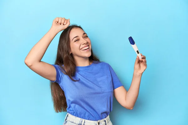 Excited woman holding a positive pregnancy test on blue studio.